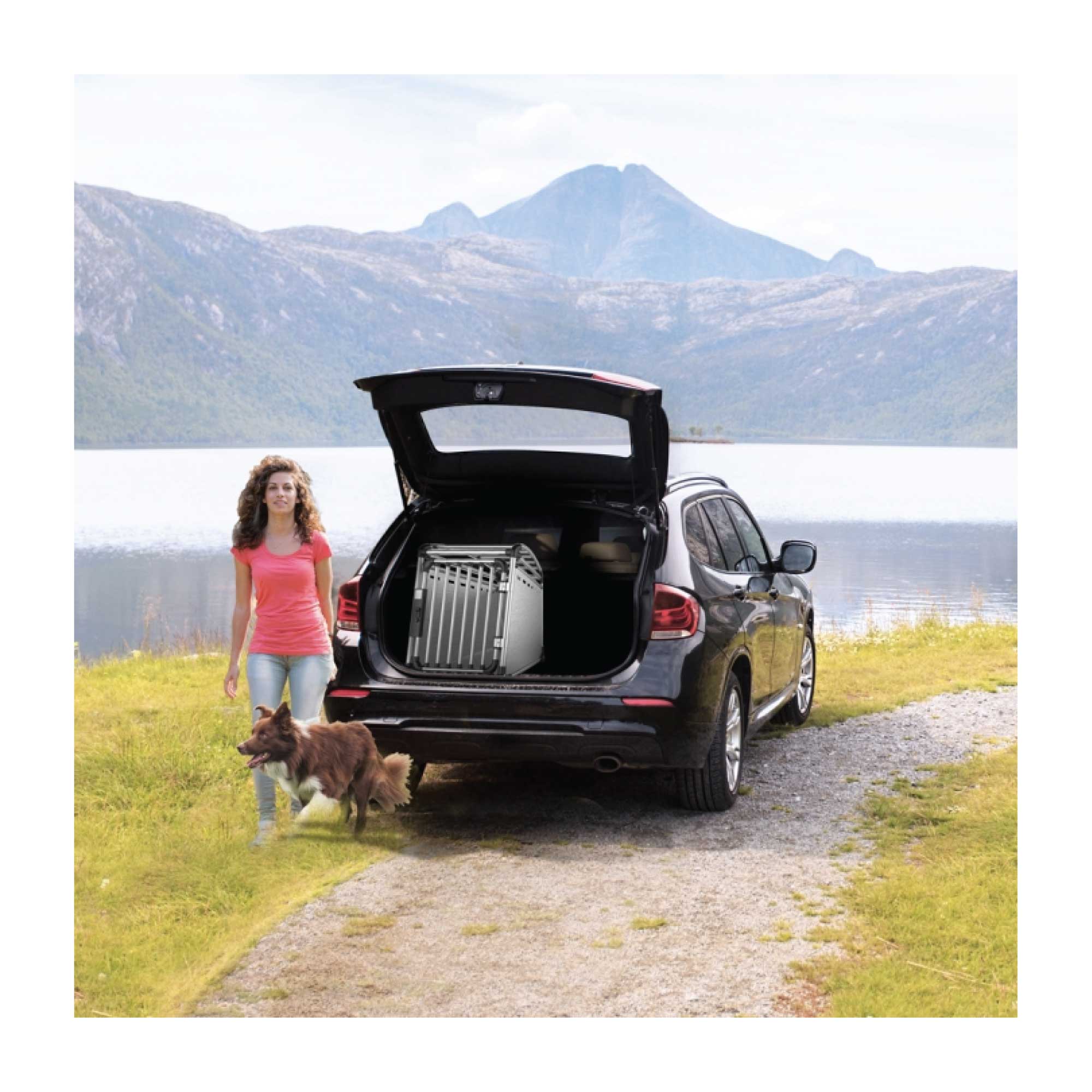 Woman with a dog walking towards an open car trunk by a lake with mountains in the background and an All For Paws aluminium dog travel crate visible in the trunk of the car. Furry Heart Paws