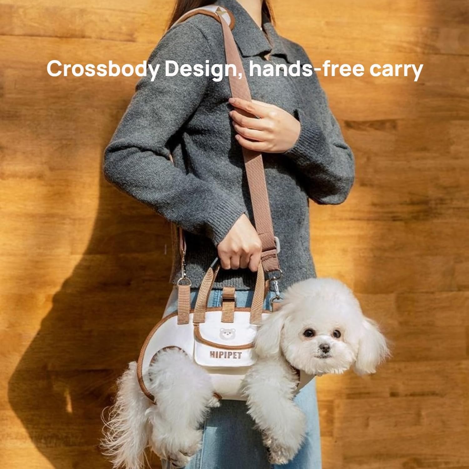 Person holding a crossbody dog carrier with a small white dog inside, against a wooden background.